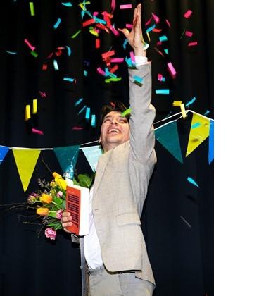 Bas Belleman, met een bos bloemen en zijn vertaling onder zijn rechterarm, steekt zijn linkerarm in de lucht. Achter hem hangen slingers tegen een zwarte achtergrond, de confetti daalt rond hem neer.